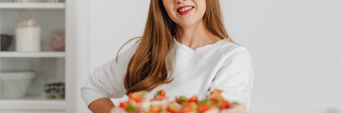 Smiling woman in a white top holding a plate of fresh canapés in a modern kitchen setting.